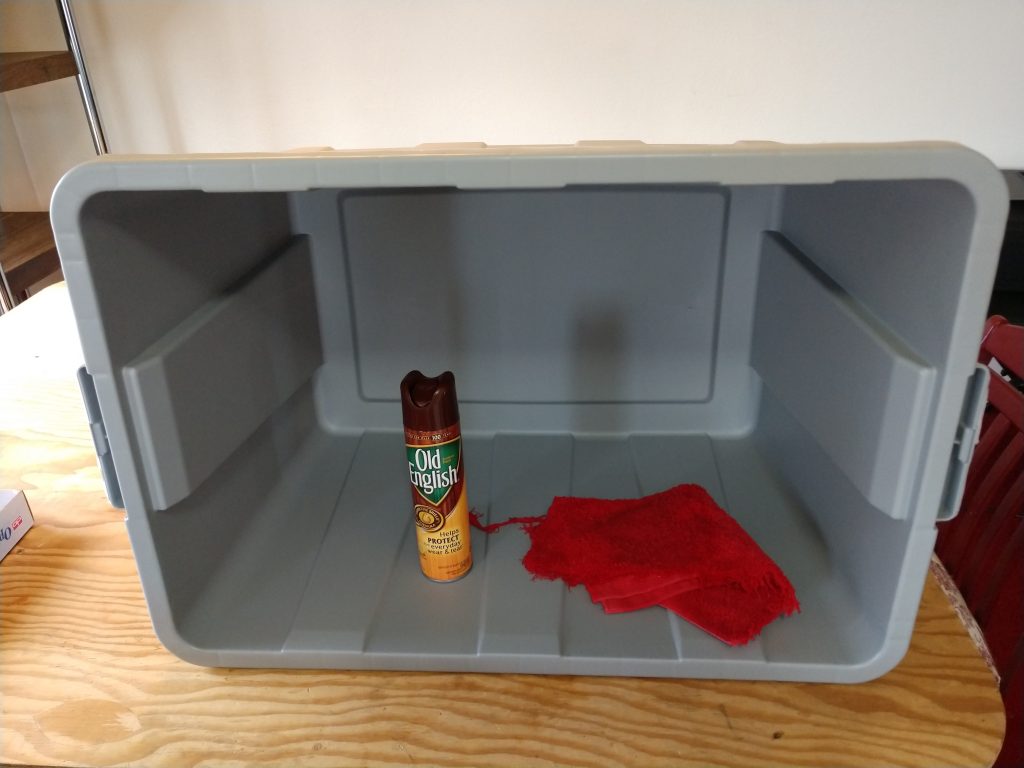 A gray storage tub laid on its side with a can of furniture polish and a polishing rag inside of the tub.